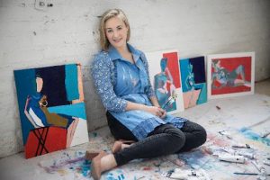 Colleen sitting on the floor in a smock. Her artwork is displayed on the floor behind her.